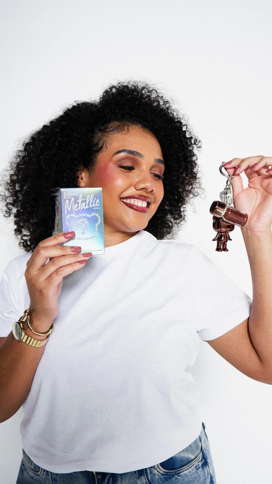 Mulher de cabelo cacheado sorrindo, de camiseta branca, segurando uma caixa holográfica em uma mão e um chaveiro de urso marrom na outra