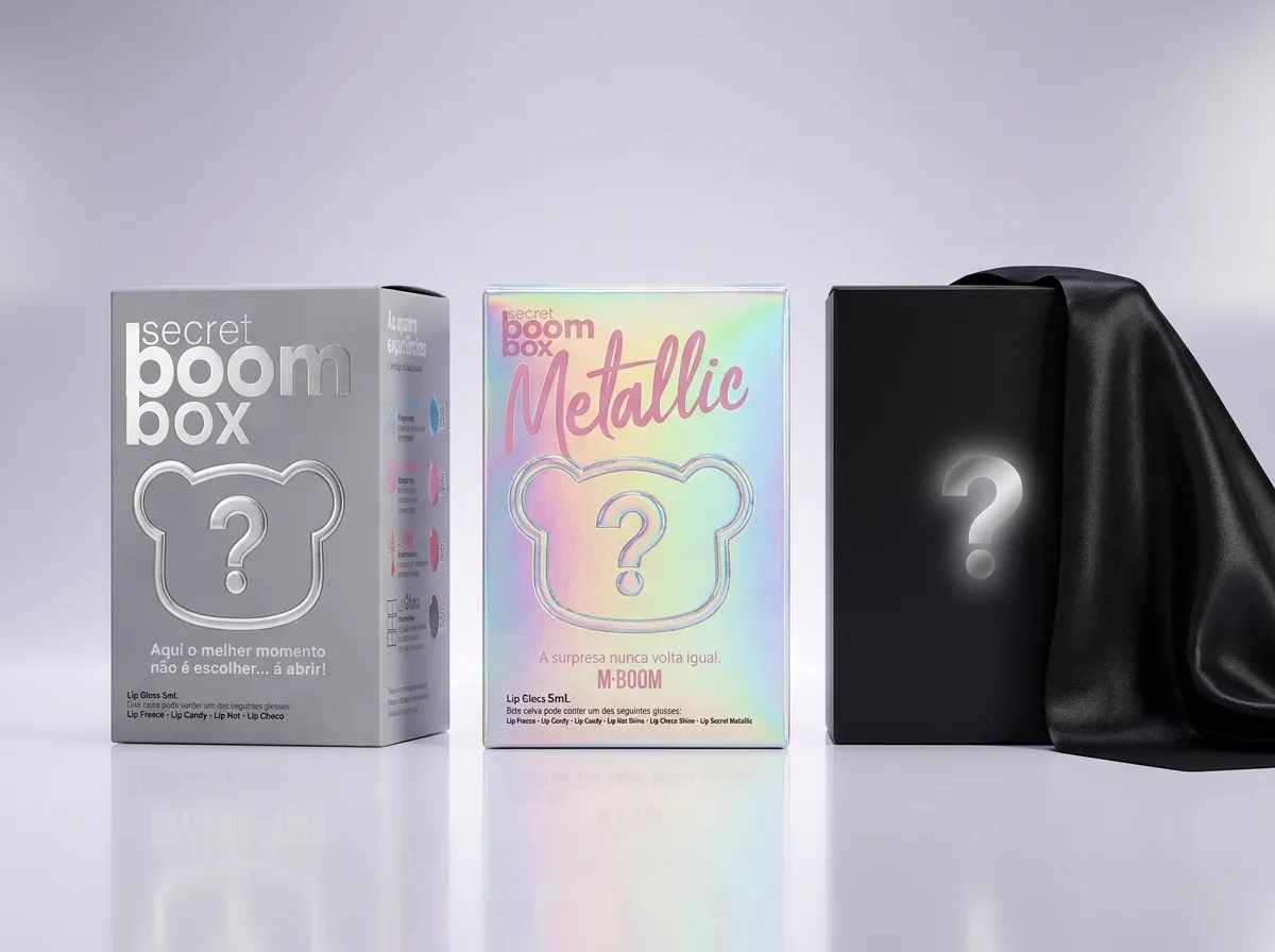 Três caixas lado a lado: a Secret Boom Box Primeira Edição cinza, a Metallic holográfica e uma caixa preta com um ponto de interrogação, coberta por um tecido de cetim